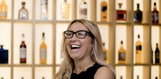 A blonde woman laughs in front of shelves full of bottles