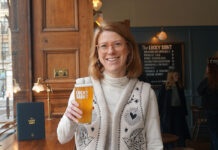A smiling blonde woman holds up a pint of beer