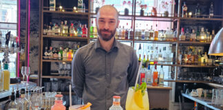 A young bearded man stands behind a well stocked bar.