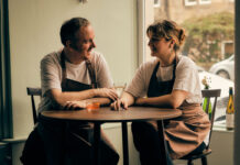 A couple wearing chefs clothes sit with drinks at a restaurant table