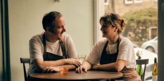 A couple wearing chefs clothes sit with drinks at a restaurant table