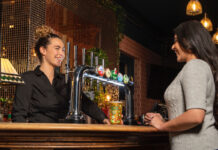 A young woman behind a bar serves another young woman a pint of cider.