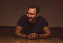 A youngish man sits at a darkly lit table smiling