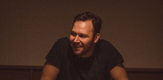 A youngish man sits at a darkly lit table smiling