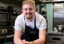 A young man with blonde hair and glasses stands in a kitchen in chef's whites