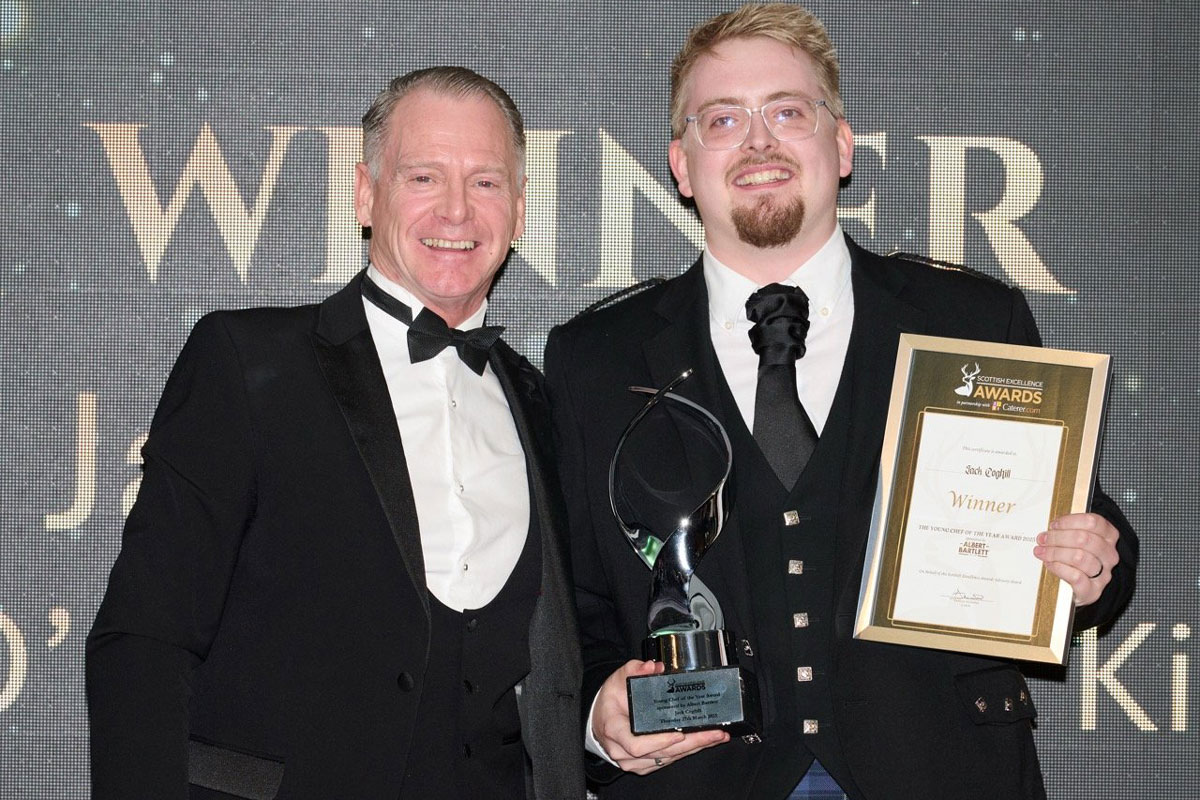 A smartly dressed young man stands on stage to receive an award from a smartly dressed older man