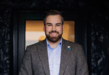 A young bearded man in a casual suit smiles at the camera