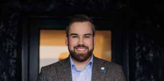 A young bearded man in a casual suit smiles at the camera