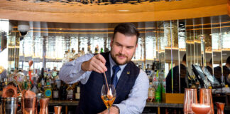 A young bearded man makes an extravagant looking drink behind a gold bar
