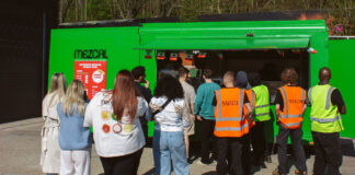 People queue at a bright green food van, in the sunshine.
