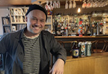 A youngish man in a beanie hat leans against a well-stocked bar