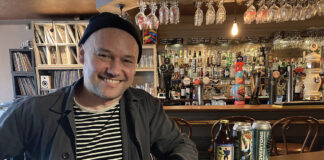 A youngish man in a beanie hat leans against a well-stocked bar
