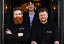 Two bearded men and a young woman sit outside a bar in matching branded clothing