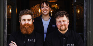 Two bearded men and a young woman sit outside a bar in matching branded clothing