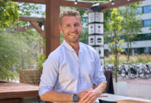 A prosperous looking blonde man stands smiling in a casual shirt