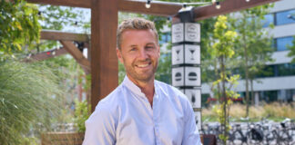 A prosperous looking blonde man stands smiling in a casual shirt