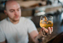 A young man in white clothes holds up a drink with a pyramidal ice cube in it