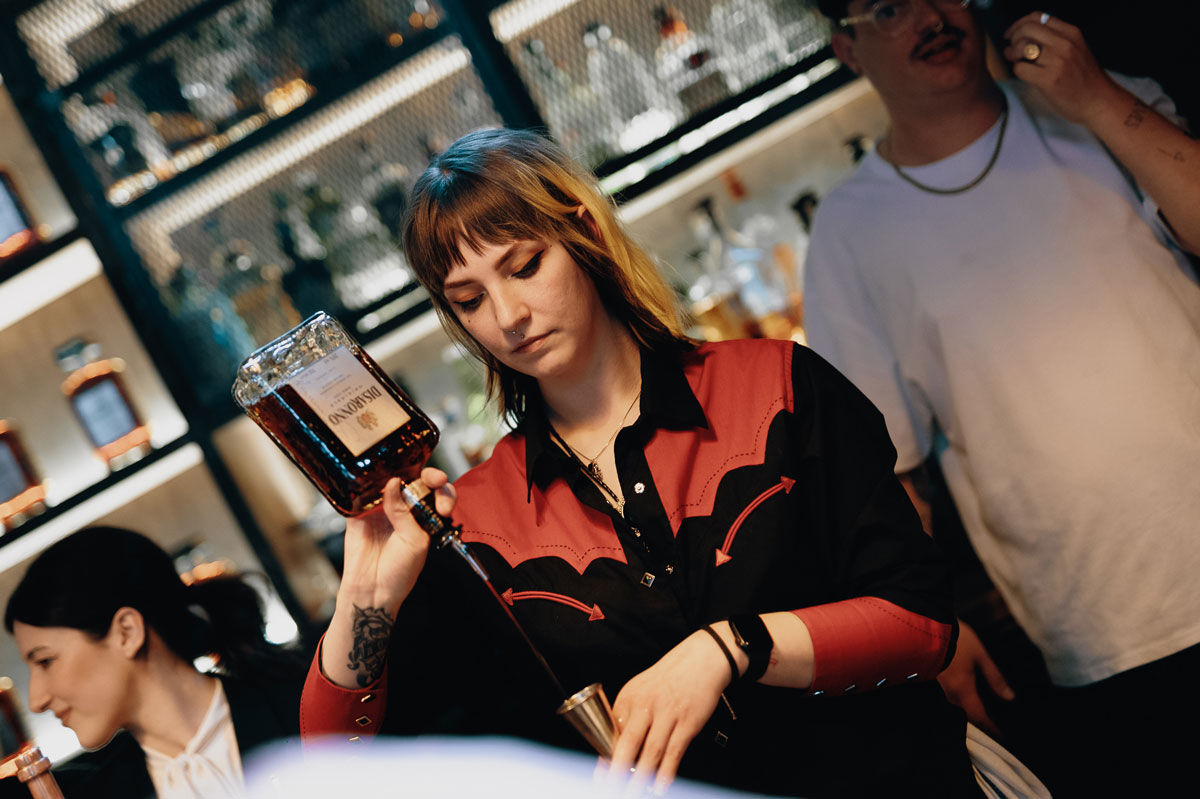 A young woman pours alcohol from a bottle into a cocktail jigger