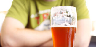 A half full pint glass sits on a table in front of a man with crossed arms