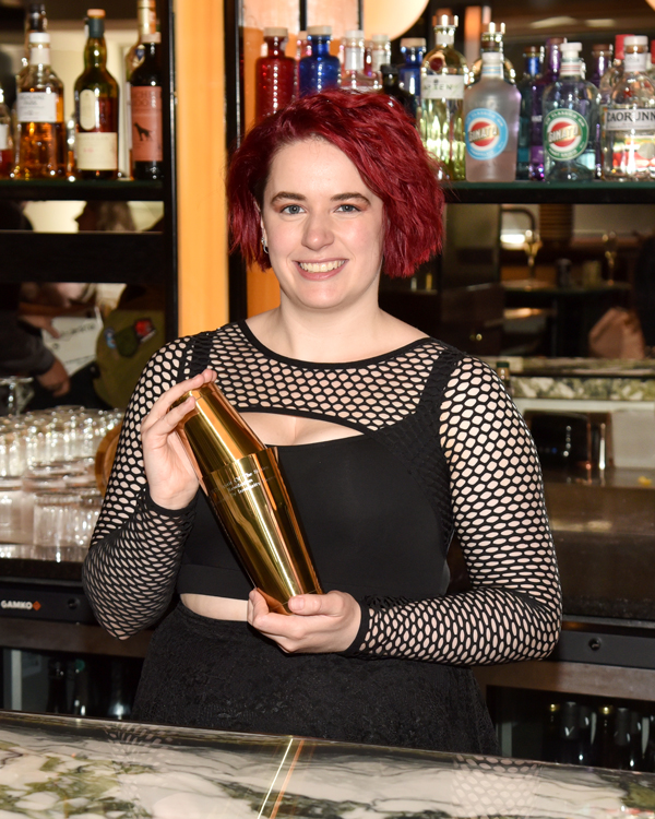 woman with red hair holding a gold cocktail shaker and smiling