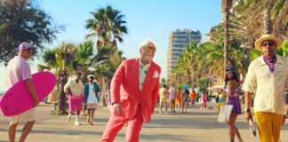 a middle-aged man in a pink suit roller-skates outside in the sunshine