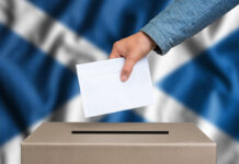 The hand of woman putting her vote in the ballot box during an election with a Scottish flag in the background.