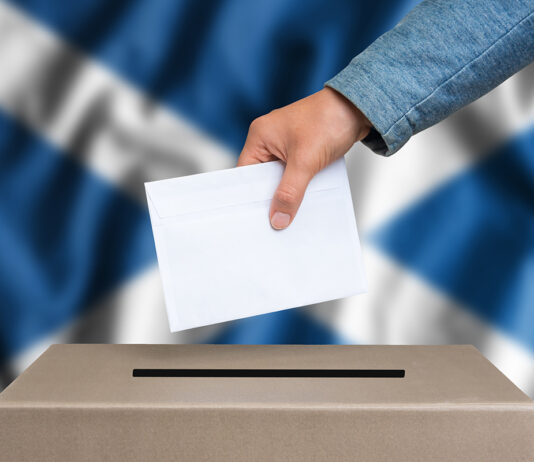 The hand of woman putting her vote in the ballot box during an election with a Scottish flag in the background.
