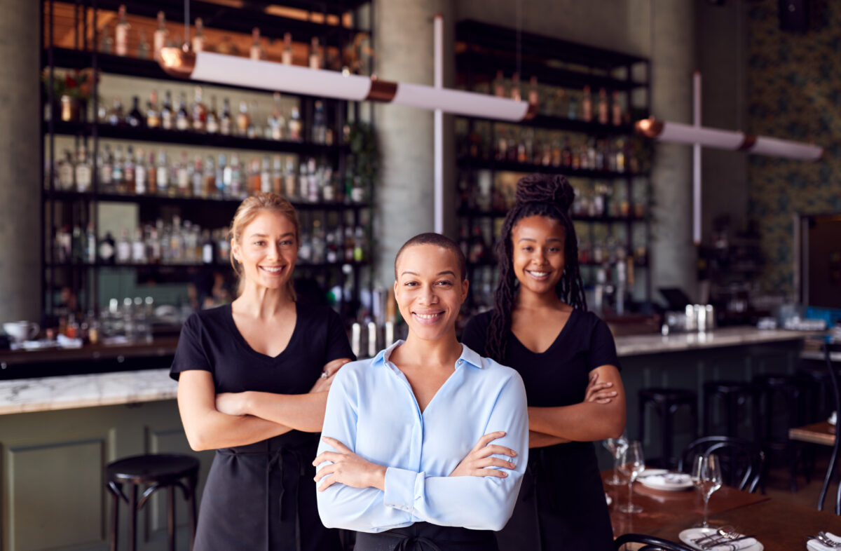 Thee female wait staff standing with arms folded and smiling in the middle of a bar/restaurant.